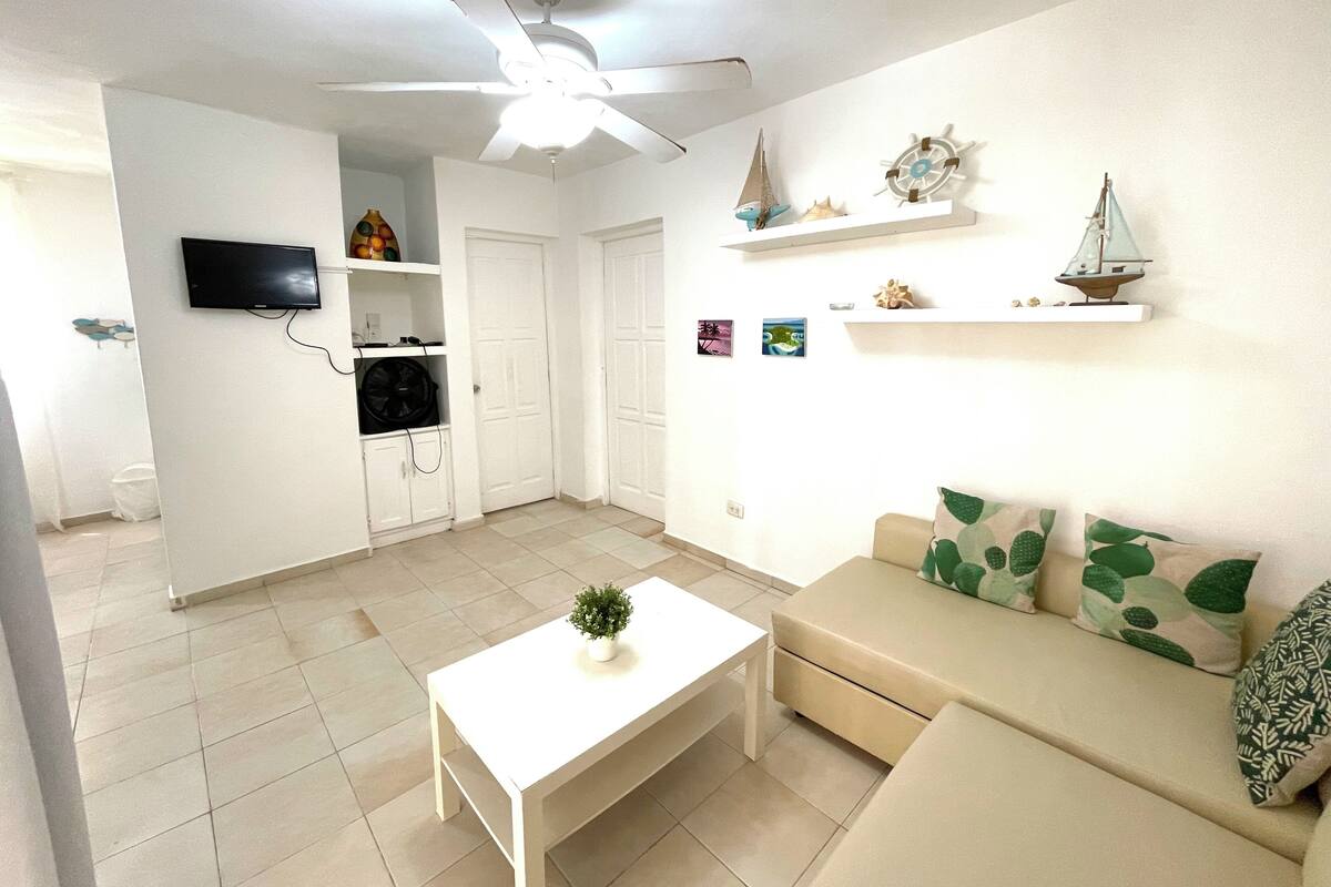 Un salon avec un canapé blanc, une table basse blanche, un ventilateur de plafond, un téléviseur fixé au mur et une petite plante sur la table.