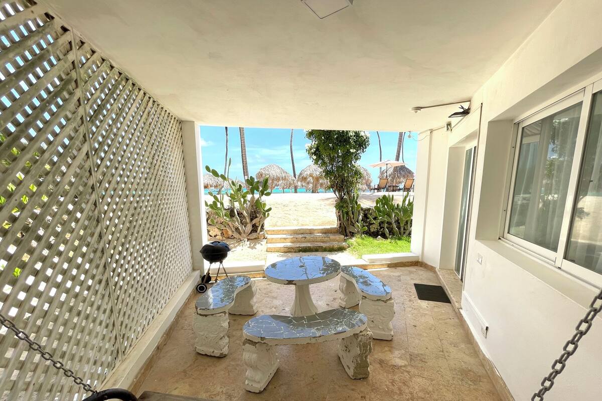 Un patio couvert avec une table et des chaises en pierre, un barbecue et une vue sur la plage et les palmiers.