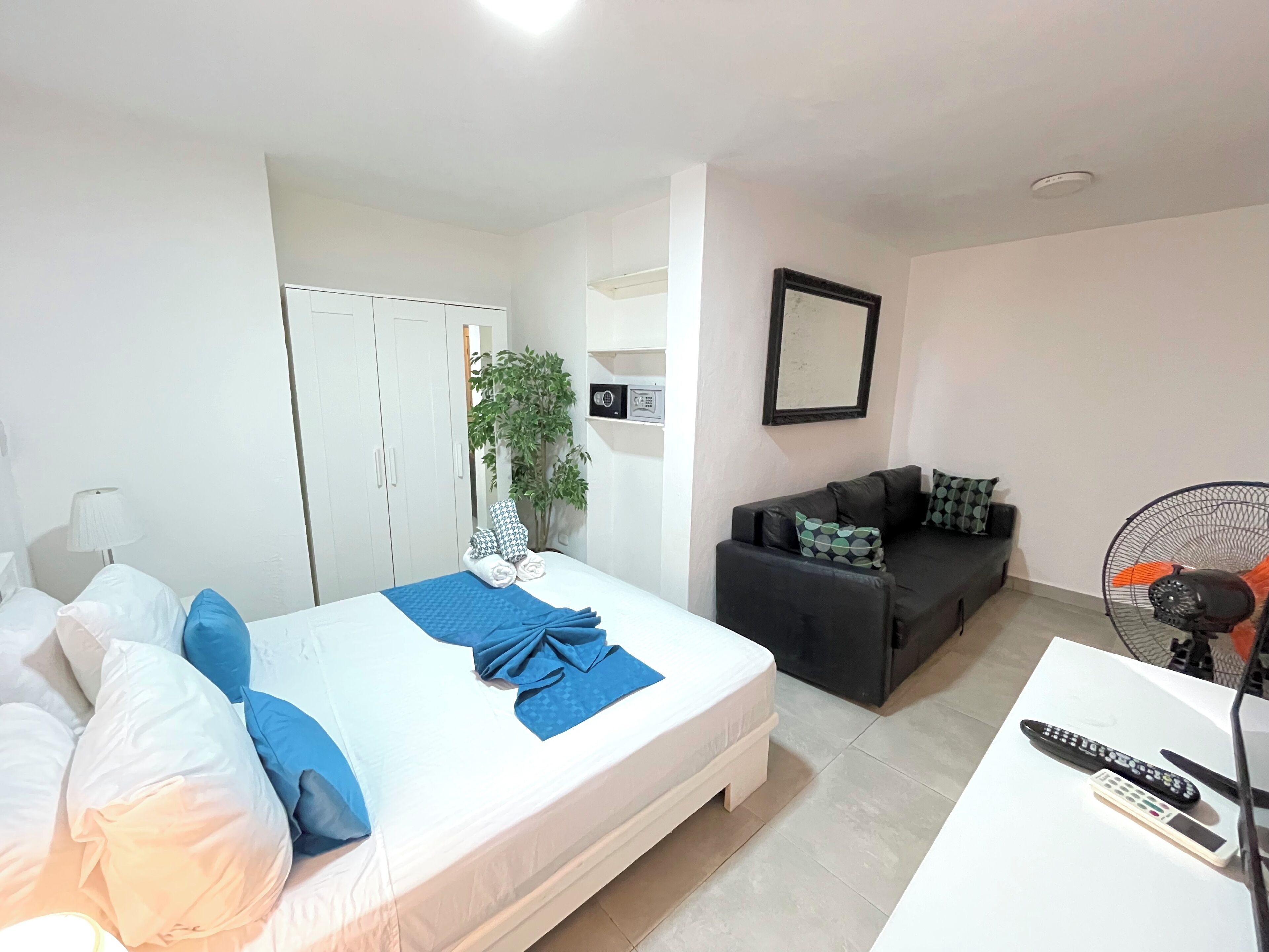 Ein Hotelzimmer mit einem Bett, einer Couch, einem Tisch mit Fernbedienungen, einer Pflanze und einem Kleiderschrank.