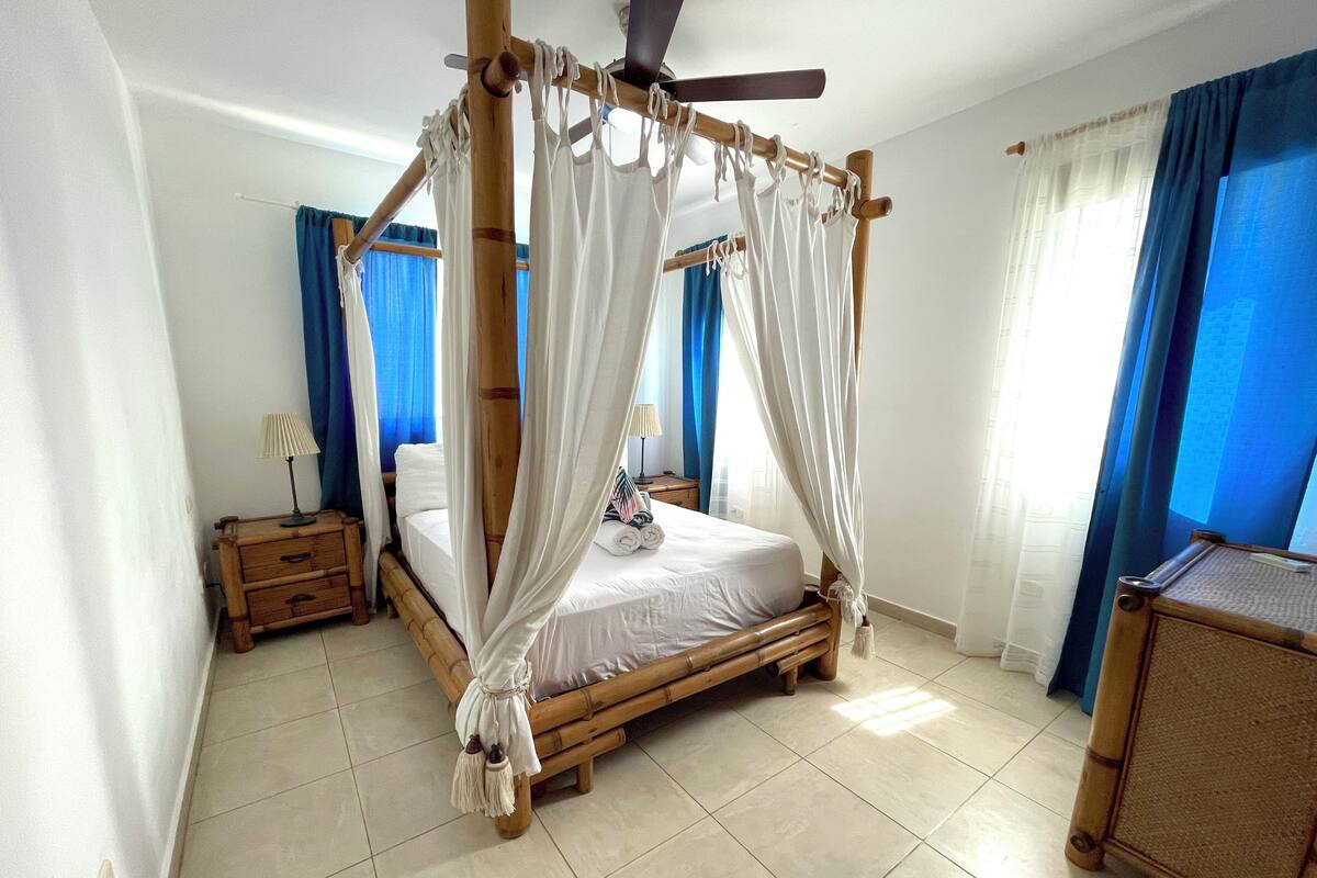 Une chambre dotée d’un lit à baldaquin, d’une table de chevet en bois, d’une petite table et d’un ventilateur de plafond.