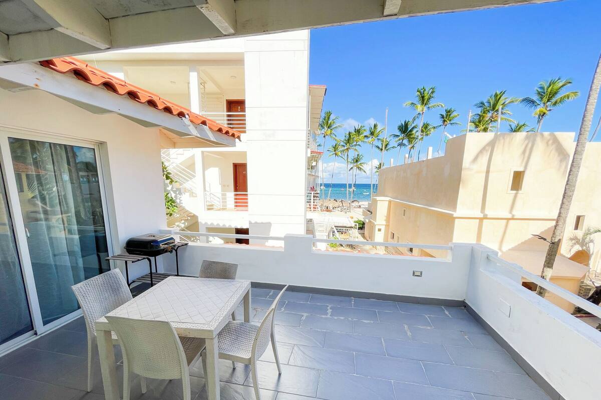 Un balcon avec une table et des chaises, donnant sur une plage bordée de palmiers et surplombant un ciel bleu azur.