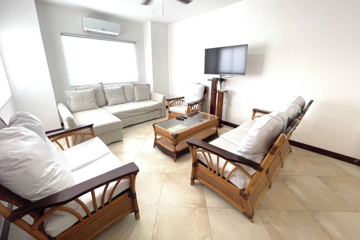 Un salon comprenant un canapé, deux fauteuils, une table basse, un téléviseur fixé au mur et un appareil de climatisation.