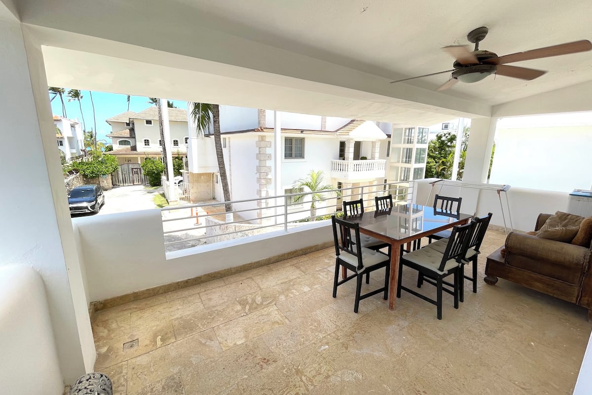 Un vaste espace de vie doté d’une grande baie vitrée donnant sur une rue résidentielle, d’un ventilateur de plafond et d’une table à manger avec des chaises.