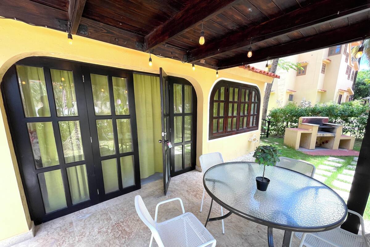 Une terrasse extérieure avec une table ronde en verre et des chaises blanches, un plafond en bois et un mur jaune agrémenté de grandes fenêtres.