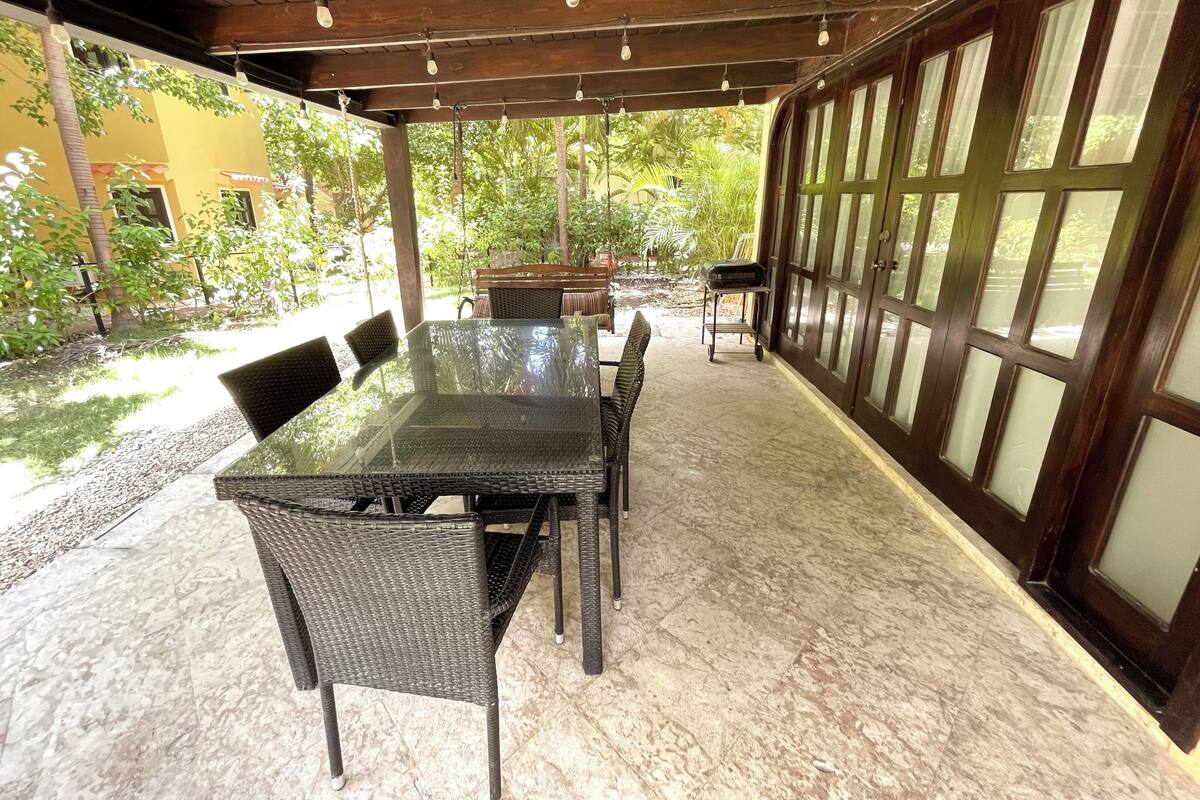 Une terrasse extérieure dotée d’une table à plateau de verre et de chaises en osier, entourée d’une pergola en bois et de grandes fenêtres.