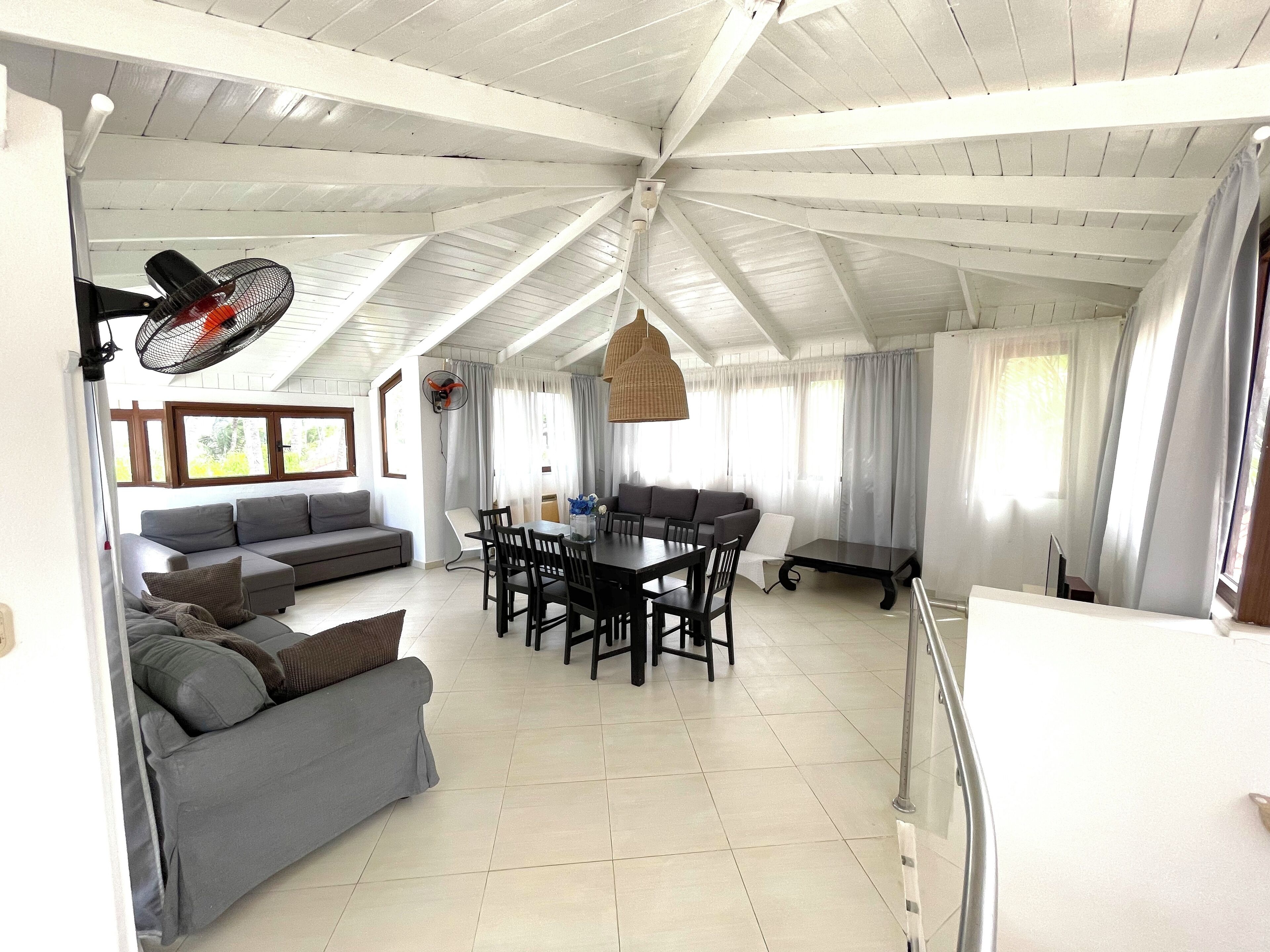 A spacious living area with a high ceiling, a dining table with chairs, a gray sofa, and a fan.
