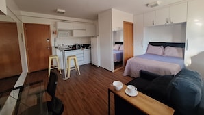 A compact hotel room with a kitchenette, a bed, a small seating area, and a glass table.