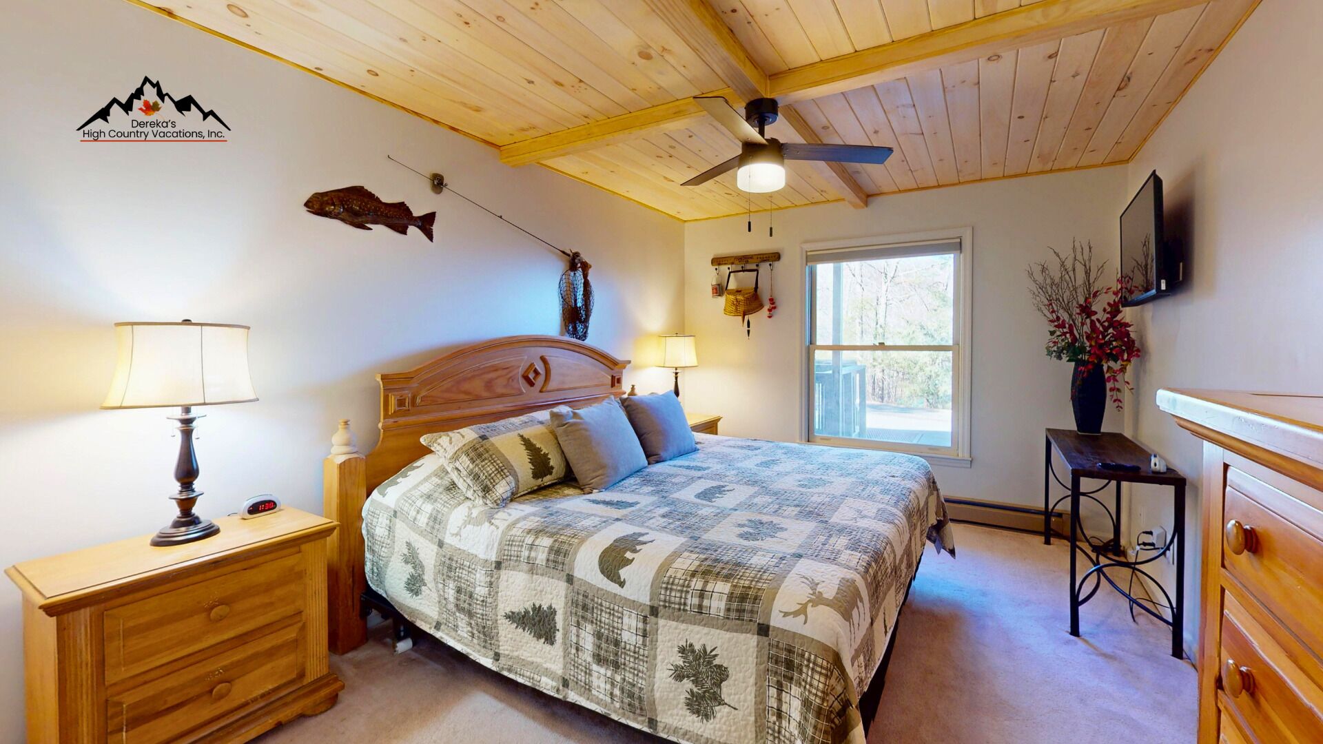 A bedroom with a wooden ceiling, a bed with a patterned quilt, two bedside tables with lamps, a wall clock, a fish decoration, and a window with a view.