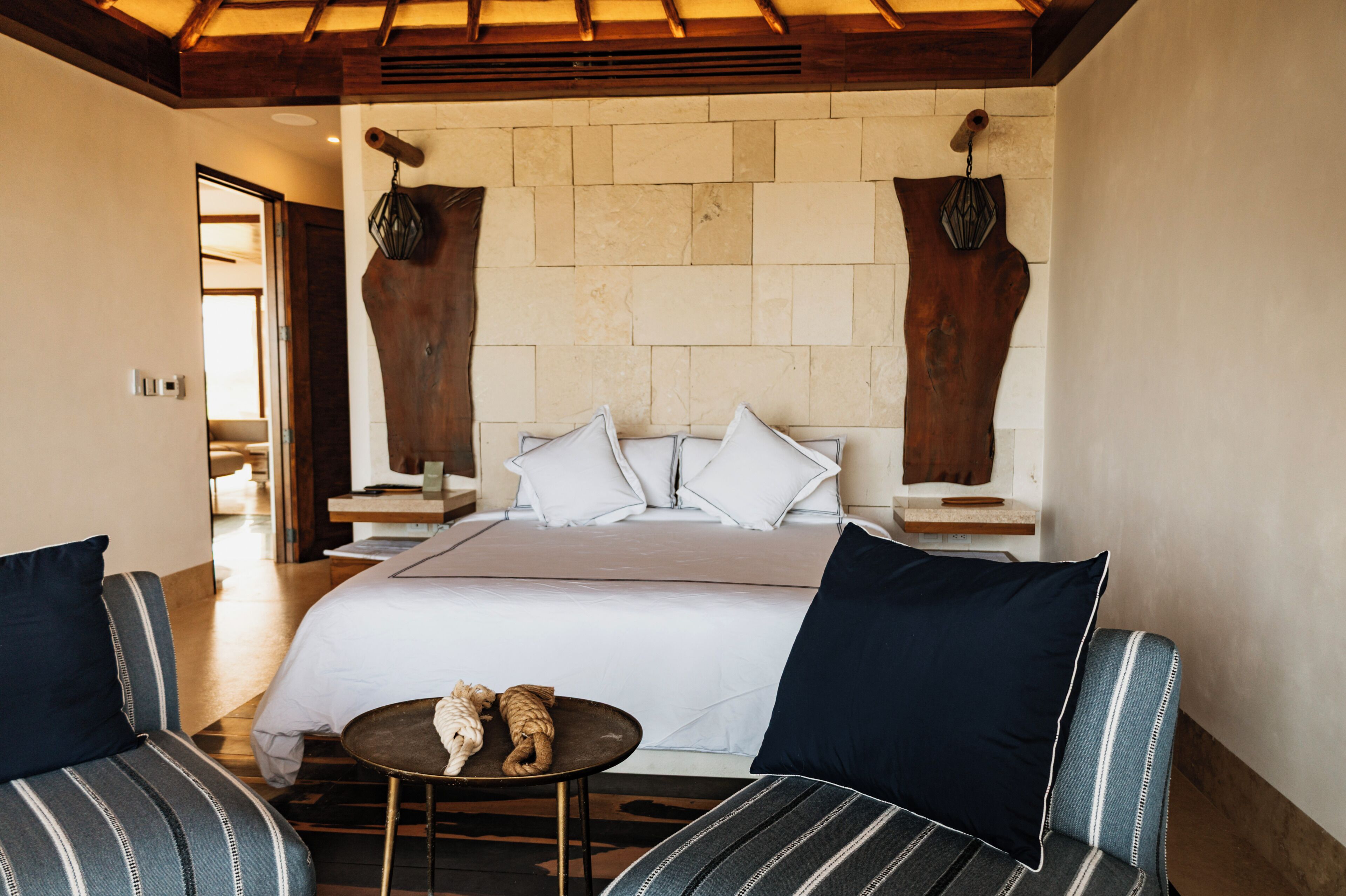 Una habitación de hotel con una cama grande, dos sillones a rayas, una mesita redonda y una pared de piedra.