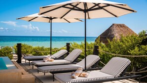 Zona junto a la piscina con sillones de descanso y una gran sombrilla, con vista al mar azul cristalino y una estructura de techo de paja.