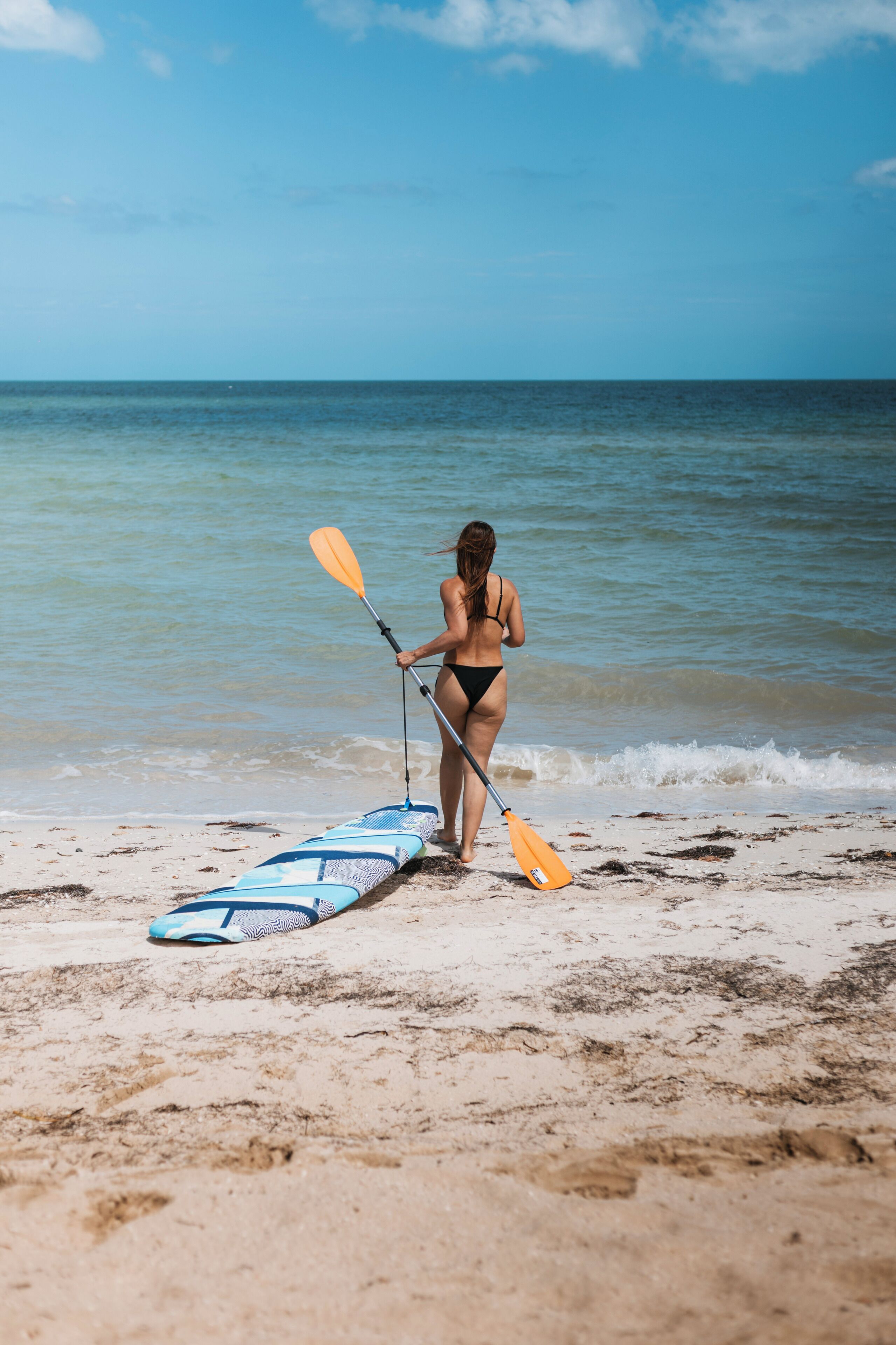 Una mujer con traje de baño de pie en una playa arenosa con una tabla de remo y un remo.