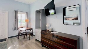 A modern room with a wooden dresser, a flat-screen TV mounted on the wall, a dining table with chairs, and a window with curtains. - Town Center Abaco (Marsh Harbour)