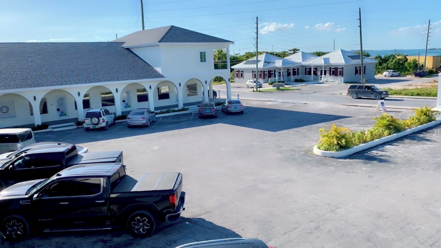 A parking lot with several cars, a building with a white facade and multiple arches, and a view of the ocean in the background.