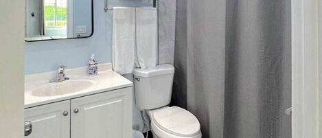 Un baño con lavamanos blanco, espejo e inodoro. Hay una cortina de ducha y un perchero con toallas.