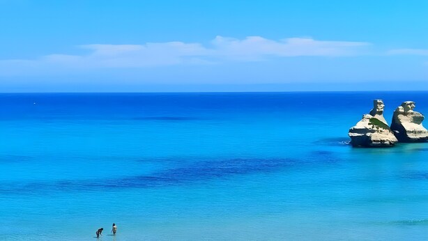 Ein Strand mit klarem Wasser, zwei Felsformationen und Menschen am Ufer.