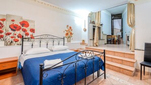 Ein Schlafzimmer mit einem Bett, einem Nachttisch, einem Bild von roten Blumen und Blick auf einen Essbereich durch eine offene Tür.