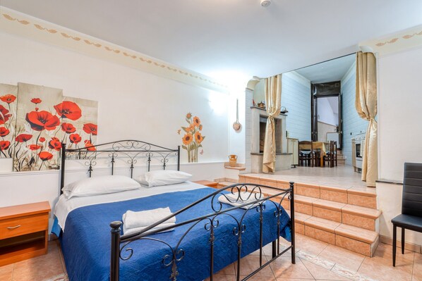 A bedroom with a bed, a nightstand, a painting of red flowers, and a view of a dining area through an open door.