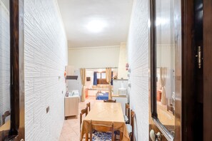 A narrow hallway with a dining table and chairs, a kitchen area with a window, and a door leading to another room.