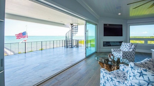 Een ruime woonkamer met een grote schuifdeur naar een balkon met uitzicht op de oceaan, een trap en een aan de muur bevestigde flatscreen-tv.