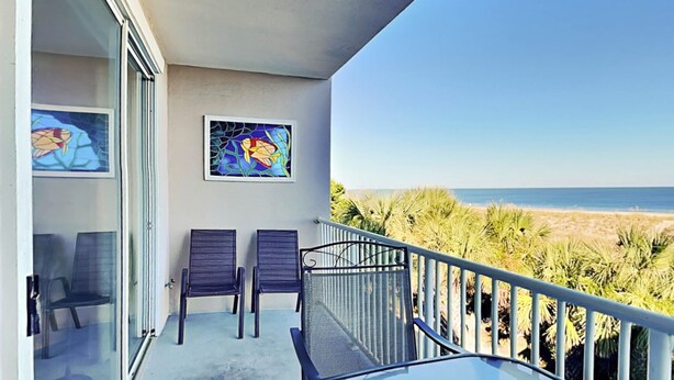 A balcony with two chairs, a glass table, and two framed artworks. The balcony overlooks a beach with palm trees and the ocean. - Ocean Song 323 | Incredible View, Two Pools (Tybee Island)