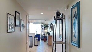 A hallway with tiled flooring, framed artwork on the walls, a potted plant, and a metal shelf unit. - Ocean Song 323 | Incredible View, Two Pools (Tybee Island)