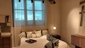 Une chambre avec une grande fenêtre, un lit avec de nombreux oreillers, une table de chevet avec une lampe et une armoire en bois.