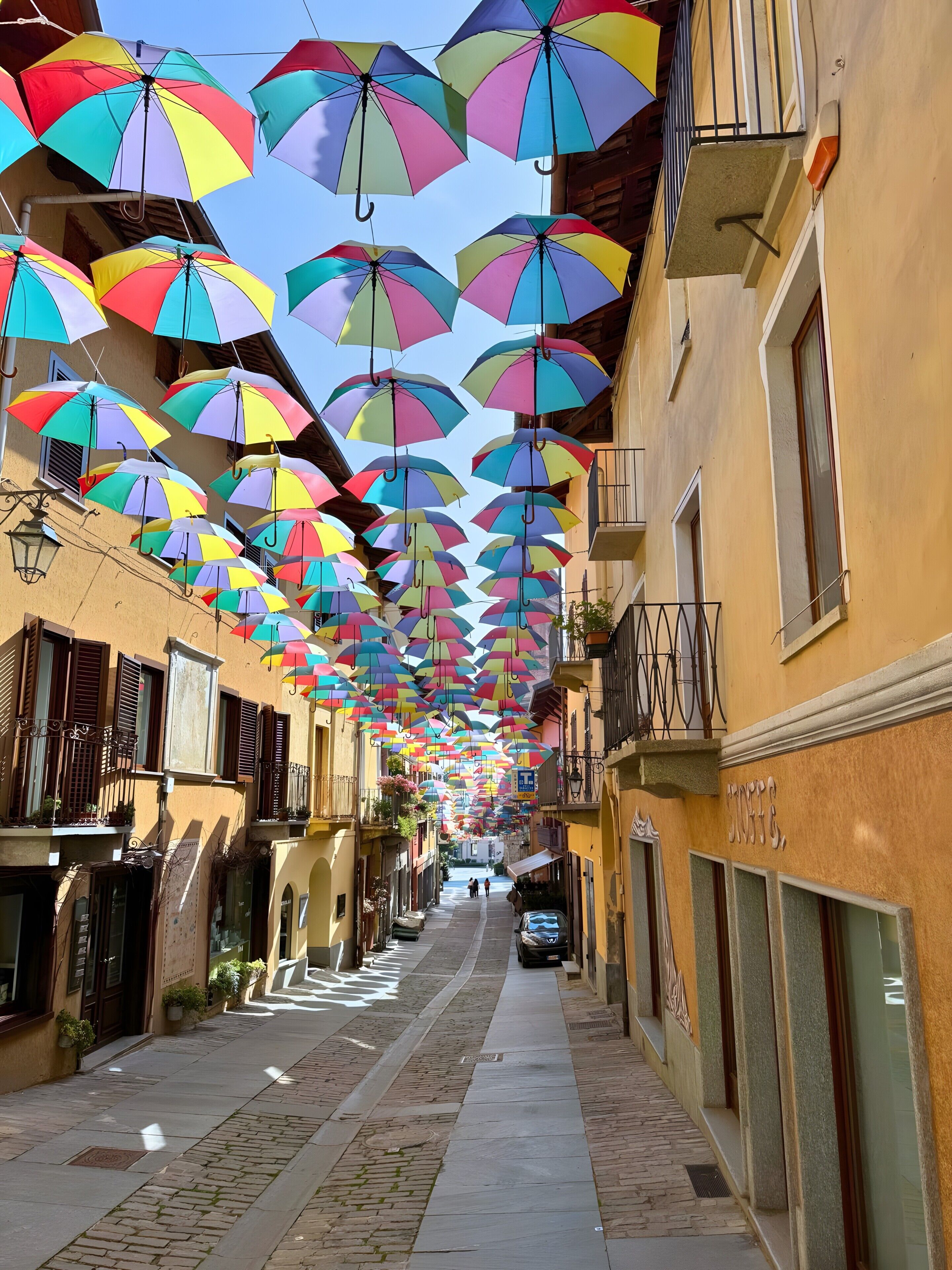 Eine enge Straße mit bunten Regenschirmen, die über dem Kopf hängen, Häuser mit Balkonen und einem klaren Himmel.