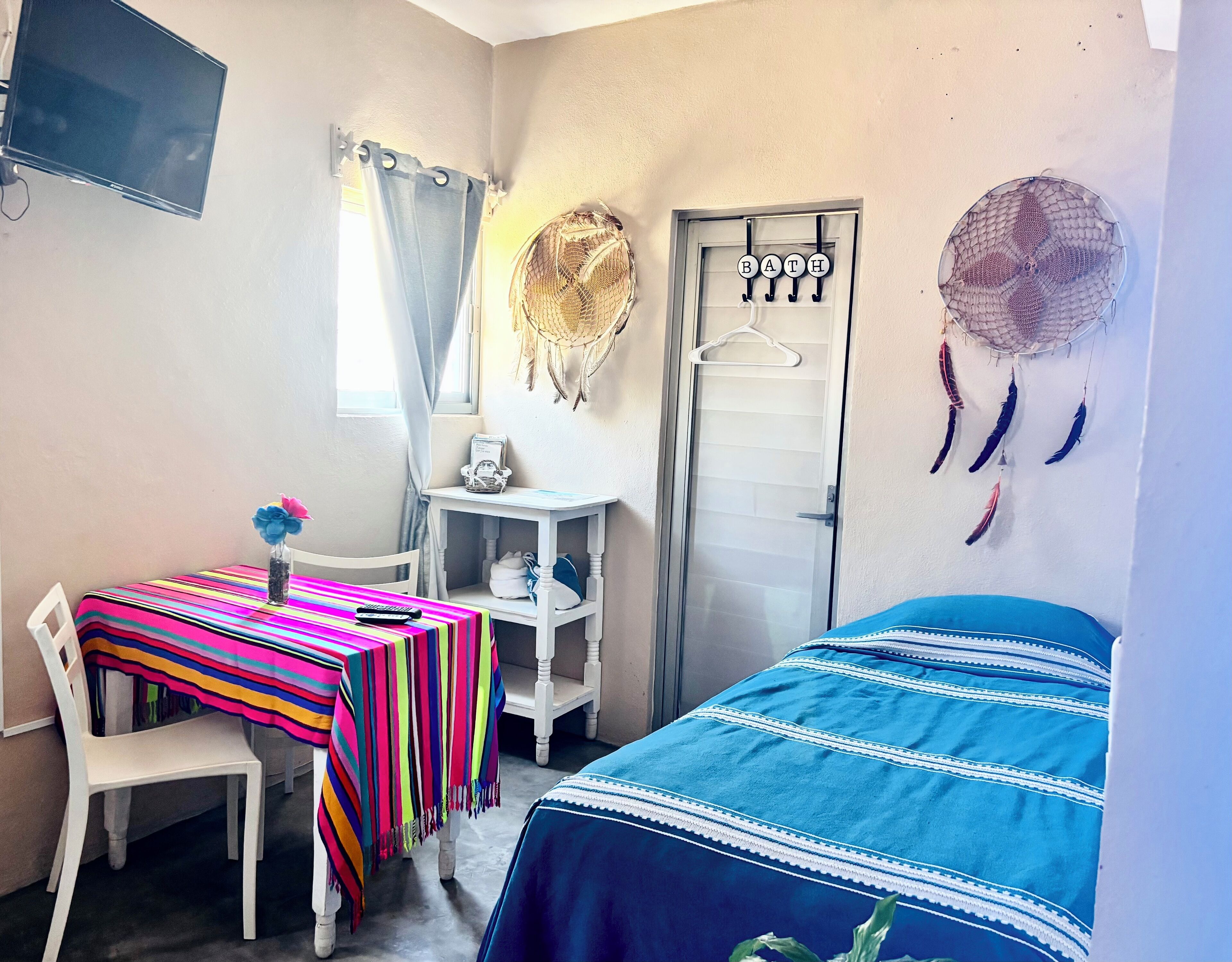 A room with a bed, a desk with a colorful striped tablecloth, a chair, a small white table with a basket, a door with a hanger, and wall decorations including a dreamcatcher and feathers.