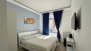 A bedroom with a single bed, a flat-screen TV mounted on the wall, a window with blue curtains, and a radiator. - Costantino Rooms (Rome)