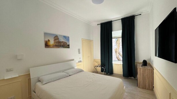 A hotel room with a bed, a painting of the Colosseum, a window with curtains, and a TV on the wall. - Costantino Rooms (Rome)
