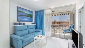 Un salon moderne avec un canapé bleu, une petite table blanche, un téléviseur à écran plat et un balcon agrémenté de chaises bleues.