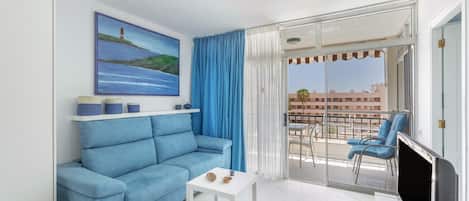 A modern living room with a blue sofa, a small white table, a flat-screen TV, and a balcony with blue chairs.