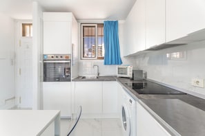 A compact kitchen with a built-in oven, a microwave, a washing machine, and a window with a blue curtain.