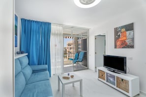 A modern living room with a sofa, a coffee table, a TV on a white cabinet, and a balcony with blue curtains.