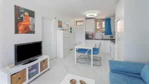 Un salon moderne doté d’un téléviseur à écran plat, d’un coin repas avec des chaises bleues, d’une cuisine avec des meubles blancs et d’un tableau accroché au mur.