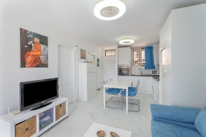 A modern living room with a flat-screen TV, a dining area with blue chairs, a kitchen with white cabinets, and a painting on the wall.