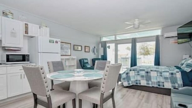 A modern, well-lit room with a round dining table, four chairs, a bed with a blue and white checkered duvet, a ceiling fan, and a view of the outdoors through large windows. - Unit 126 Beautiful Cedar key (Cedar Key)