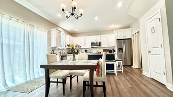 Una cocina moderna con armarios blancos, un refrigerador de acero inoxidable y un comedor con mesa y sillas de madera.