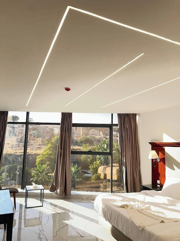 A modern hotel room with a large bed, a desk, and a view of a natural landscape through large windows. - Jerash Hotel (Jerash)