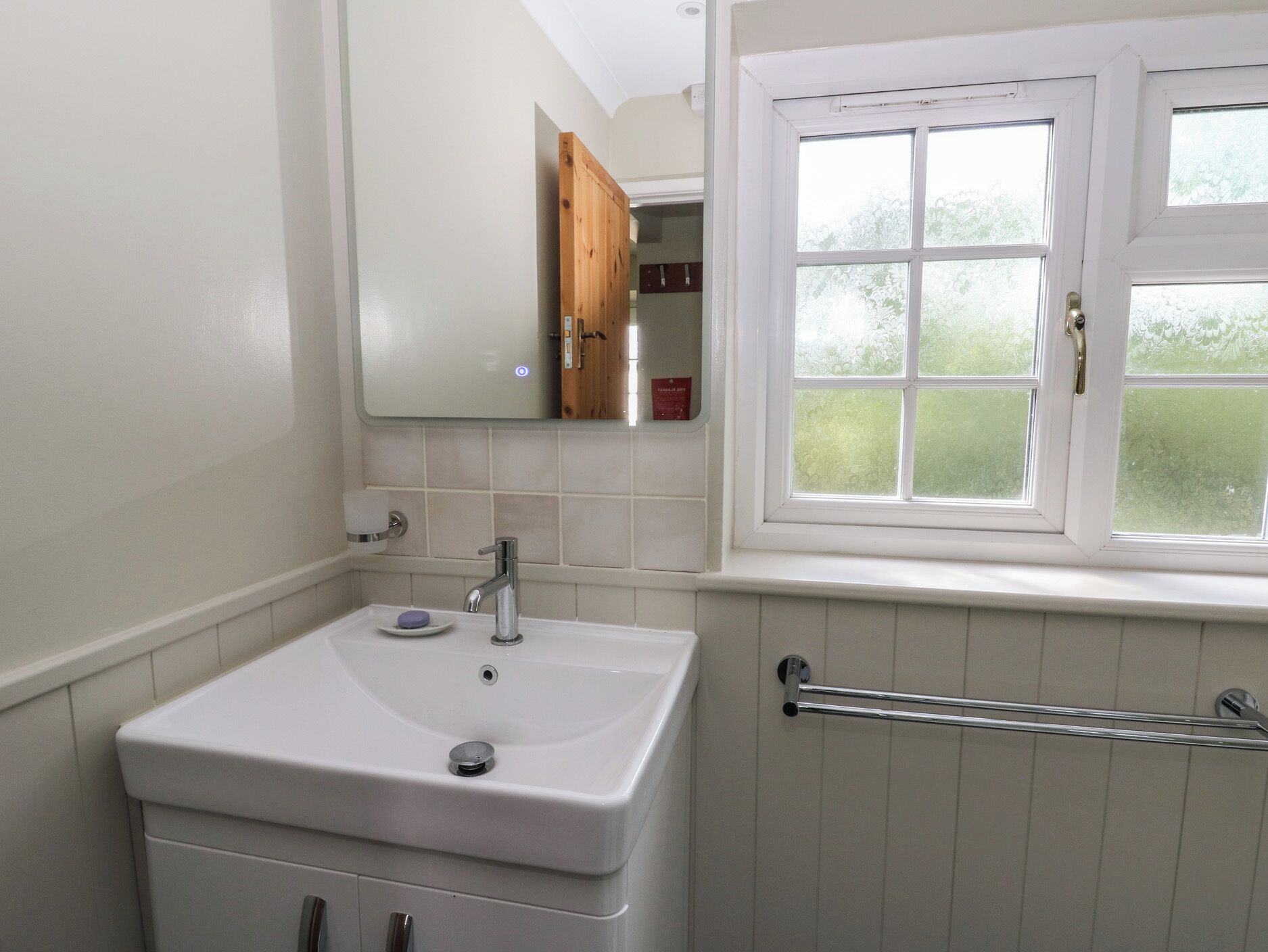 Un baño con un lavamanos blanco, un espejo, una ventana y un perchero para toallas.