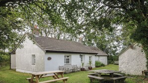 A rustic white cottage with a sloped roof, a chimney, and a small porch with two wooden benches. - Llainorfa (New Quay)