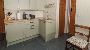 A compact kitchen with light green cabinets, a wooden countertop, and a small sink. There is a chair with a patterned cushion and a wooden door in the background. - Llainorfa (New Quay)