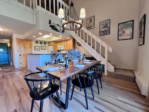 A dining area with a wooden table and chairs, a staircase, and kitchen in the background.