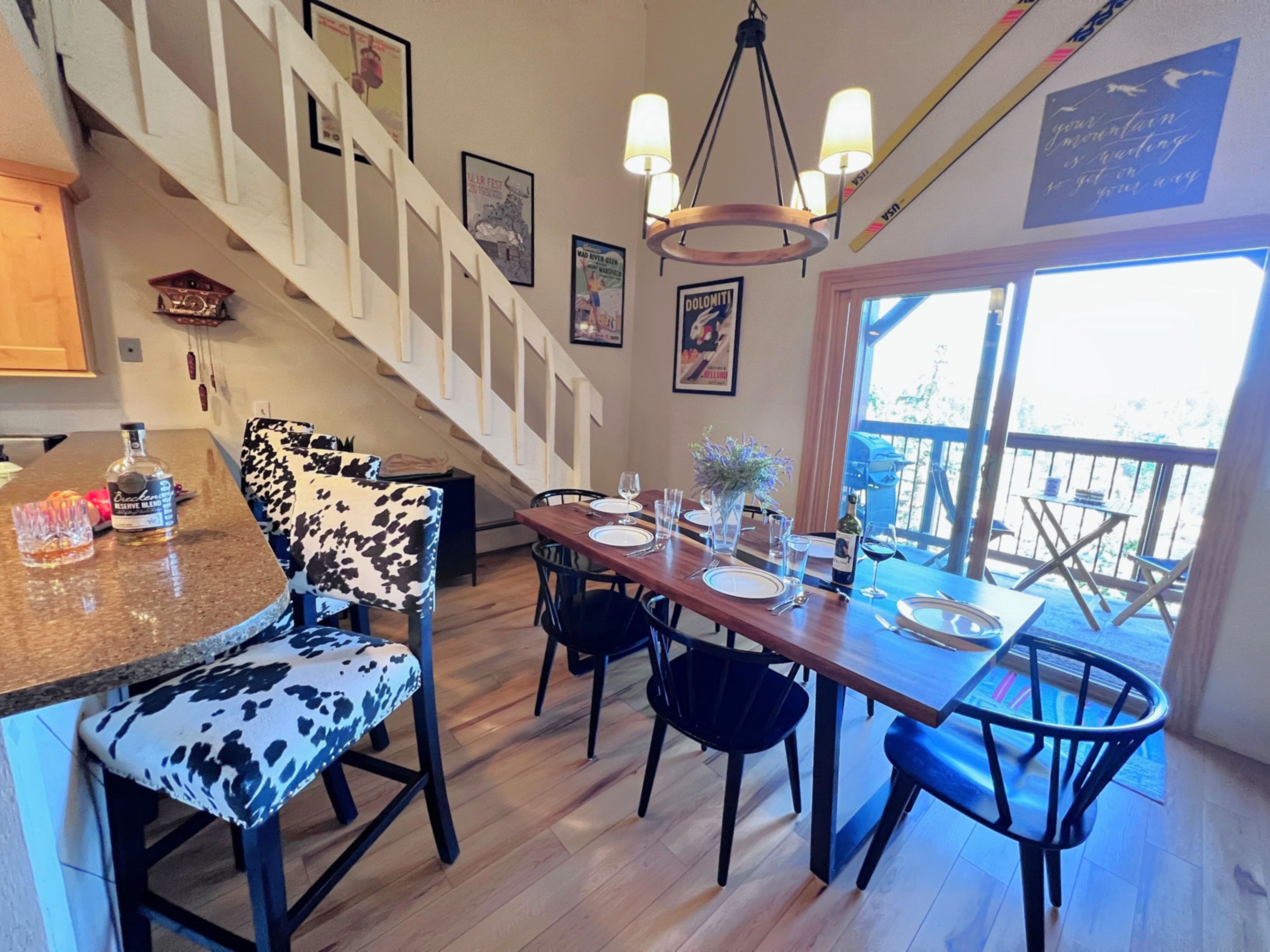 Un coin repas avec une table en bois dressée pour quatre personnes, entourée de chaises dont le revêtement est orné d’un motif noir et blanc. Un escalier avec des rampes blanches mène à un niveau supérieur.