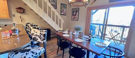 Un coin repas avec une table en bois dressée pour quatre personnes, entourée de chaises dont le revêtement est orné d’un motif noir et blanc. Un escalier avec des rampes blanches mène à un niveau supérieur.