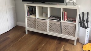 En stue med et fladskærms-tv på et hvidt tv-møbel, en hvid aircondition ovenover, og en reol med bøger og pynteting.