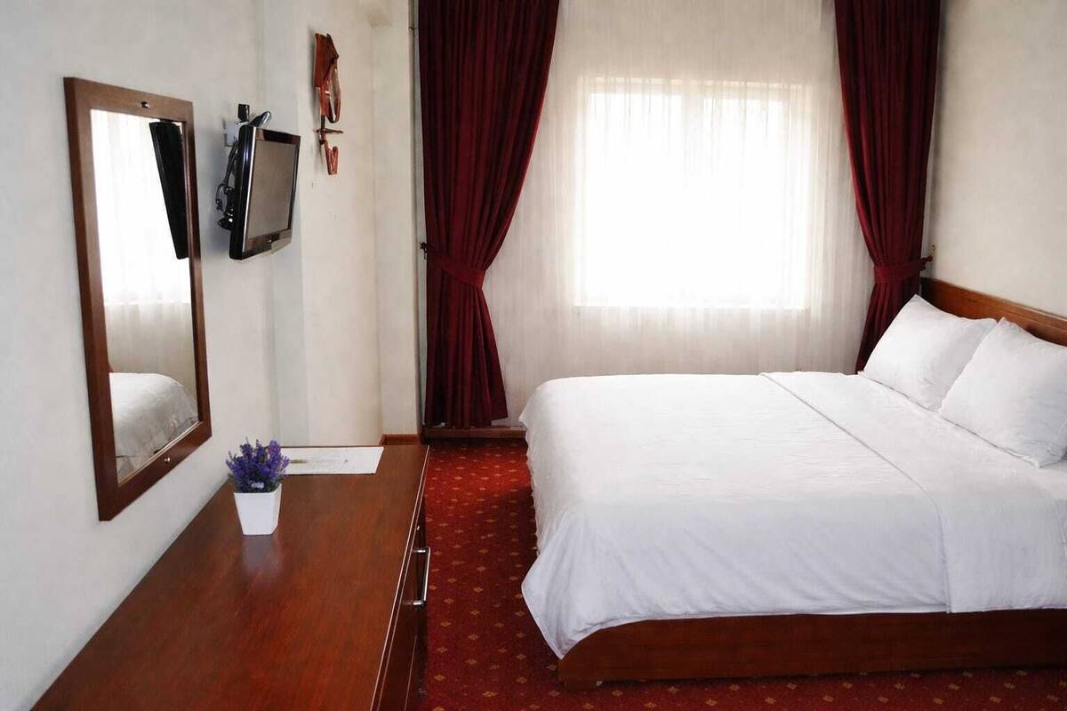 Une chambre d’hôtel avec un grand lit, une table de chevet ornée d’un vase de fleurs, un téléviseur fixé au mur et un grand miroir.