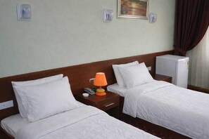 A hotel room with two beds, a nightstand with a lamp, a refrigerator, and a painting on the wall.