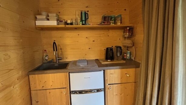 A compact wooden kitchen with a refrigerator, sink, and shelves with kitchenware. - COSY STUDIO IN A QUIET LOCATION. NEAR A TUBE STATION. (Pinner)