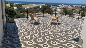 A balcony with patterned floor tiles, two wicker chairs, and a view of the sea and buildings.